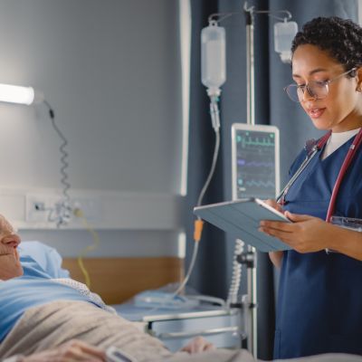 Hospital Ward: Friendly Head Nurse Talks with Elderly Patient Resting in Bed. Physician Uses Tablet Computer, Does Checkup, Ask Health Care Questions. Old Man Fully Recovering after Successful Surgery